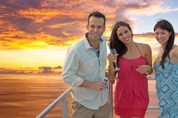 A man with a beer and two women with drink at the edge of the deck on the Big Sunset Cruise with the sunset in the background