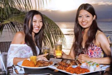 Two women enjoying BBQ and beer beachside while the sun sets over the ocean in the background in Tumon, Guam